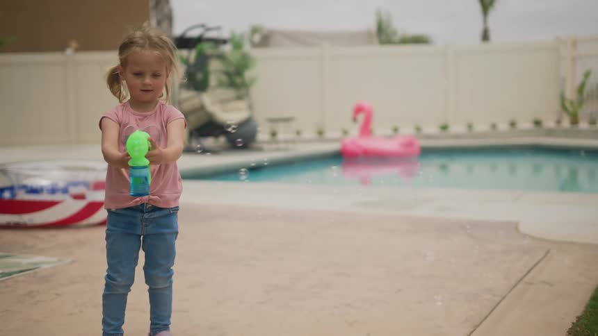 Little girl playing with a toy bubble gun near a swimming pool in the backyard