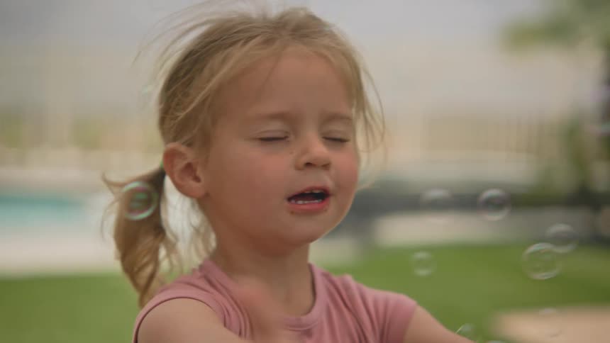 The face of a happy little blonde girl who is blinking because of soap bubbles that are heading towards her