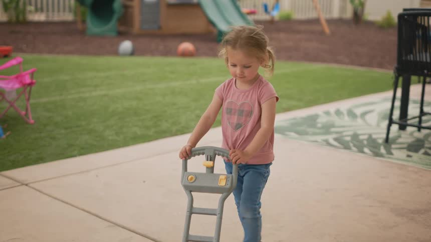A young child pushes a toy lawnmower across a paved area
