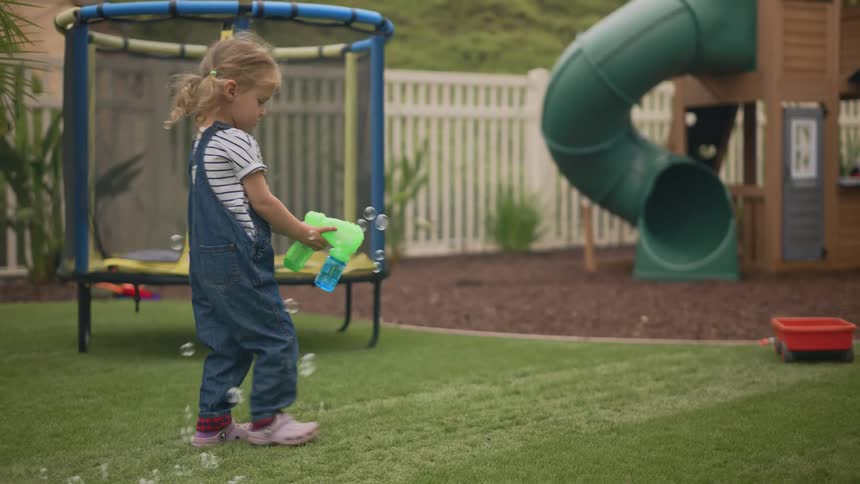 A young girl with pigtails wearing denim overalls is standing on a lawn using a bubble gun