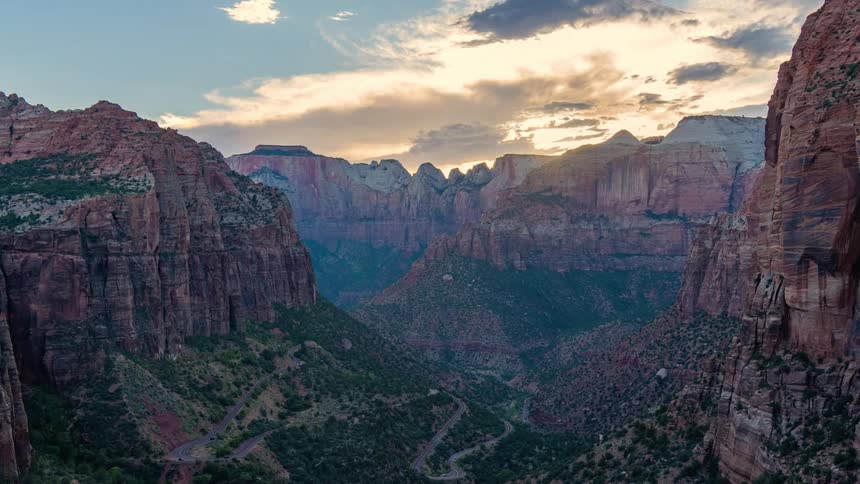 A view of a zion park with towering rock formations a winding road and a sunset illuminating the sky with clouds