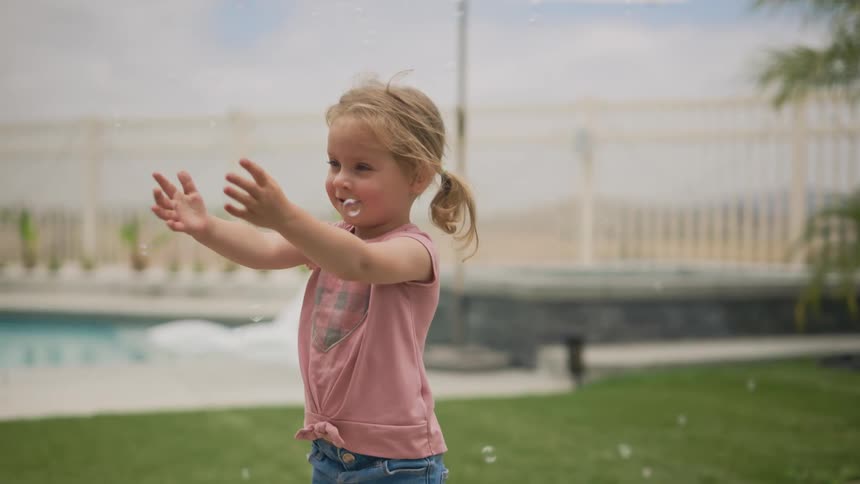 A young girl with pigtails initially reaches out then covers her eyes before delightedly interacting with bubbles in her backyard eventually picking up an object