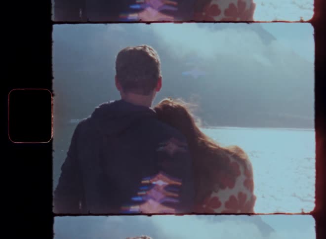 A man and a woman in coats are standing with their backs turned and the woman leans her head on the mans shoulder and they are looking out to sea