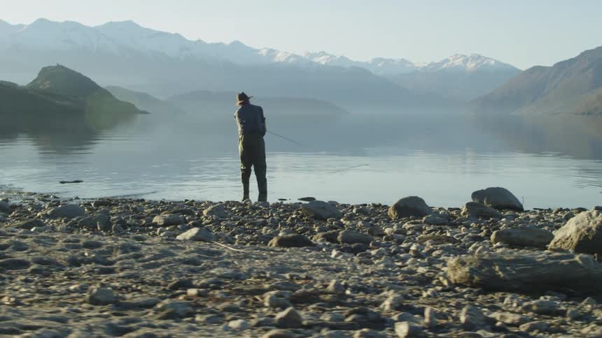 Man standing on rocks by the shore fly fishing on a cloudy day