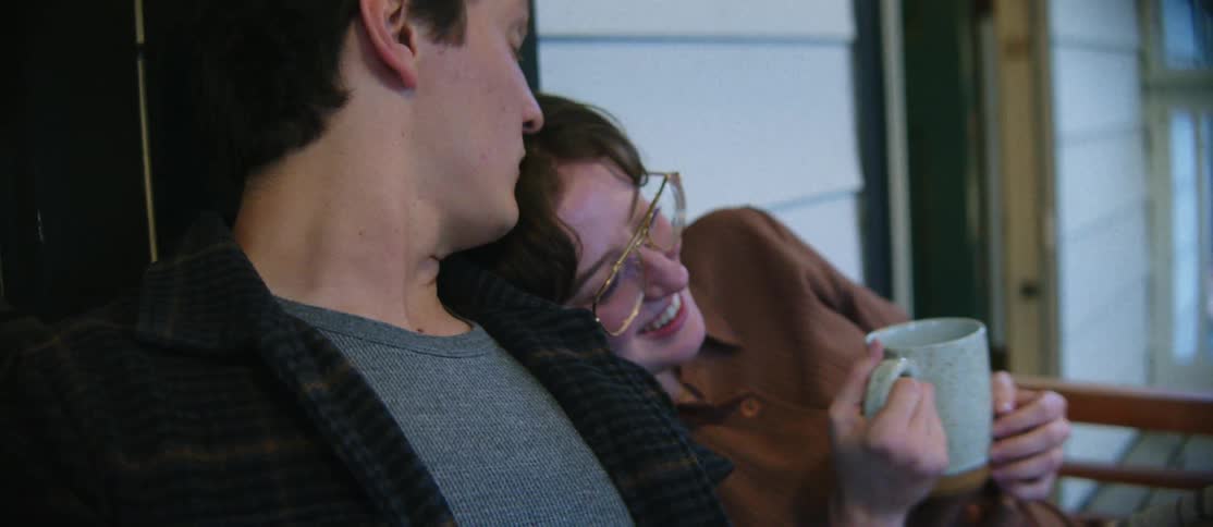 Two people are sitting closely together on a porch with one person holding a mug and leaning affectionately on the other