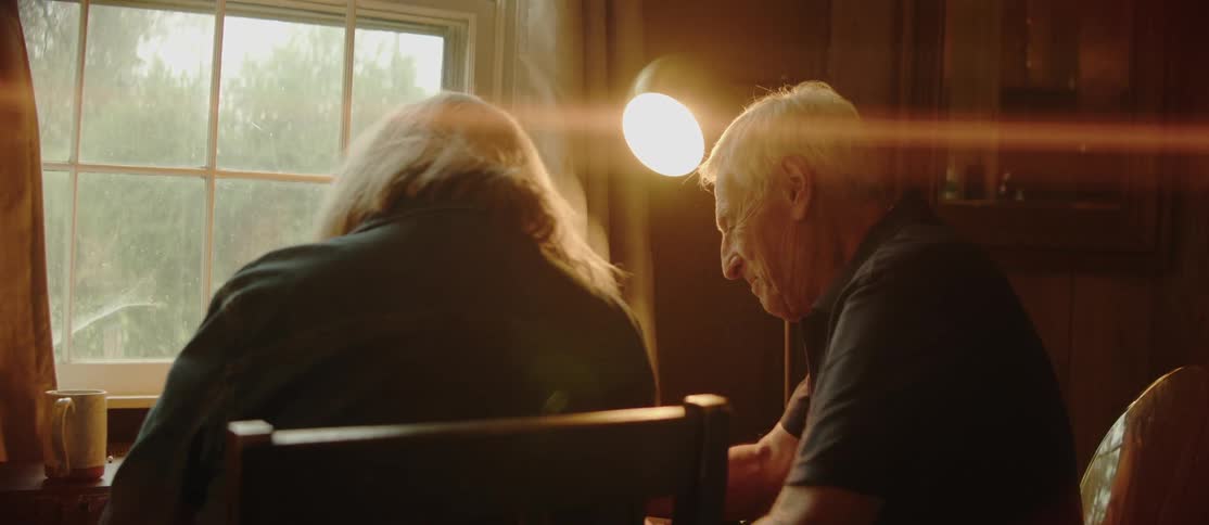 Two people are sitting at a table near a window playing music on a musical keyboard with a lamp illuminating their workspace