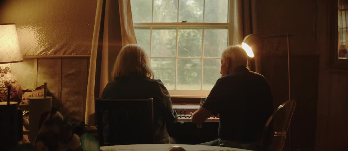 An elderly man and woman are sitting back to back in chairs and talking while the man plays the piano