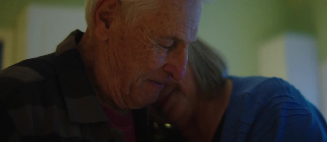 Two elderly individuals are leaning close to each other dancing in a warmly lit indoor setting