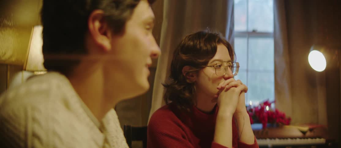 A young woman with glasses sits with her hands clasped listening intently as a young man beside her speaks
