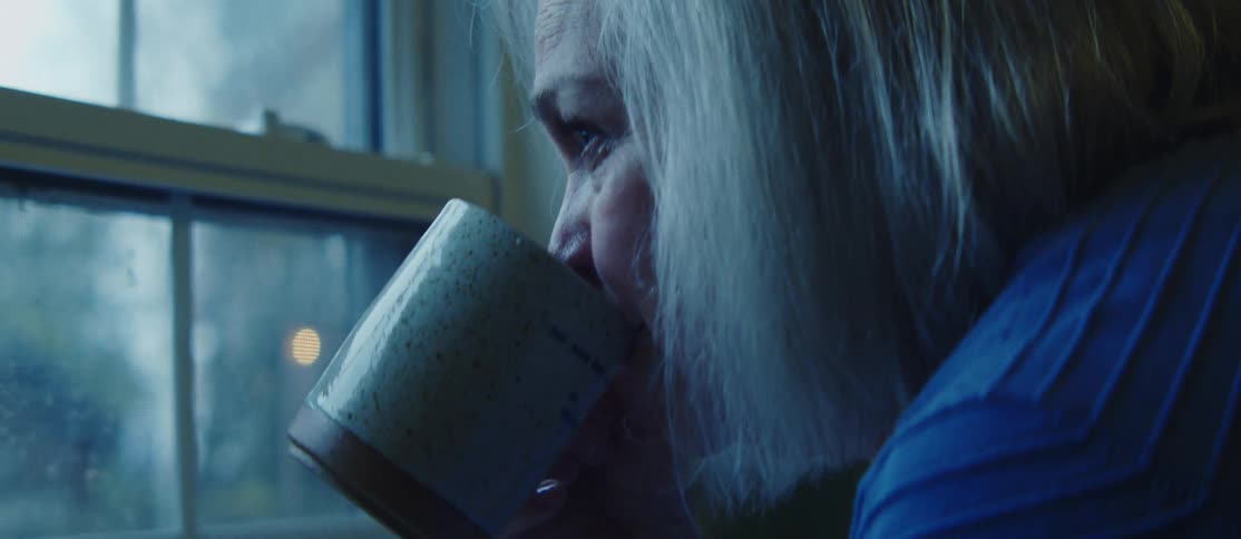 An elderly woman with long lightcolored hair is drinking from a speckled ceramic mug while looking out of a window