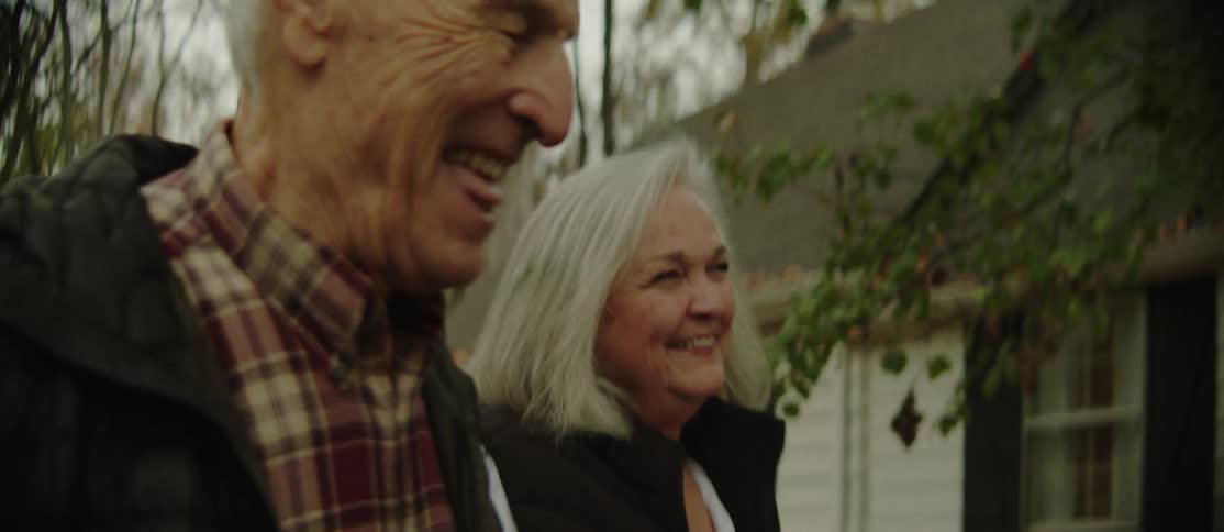 An elderly man and woman trek through the countryside passing a charming cottage