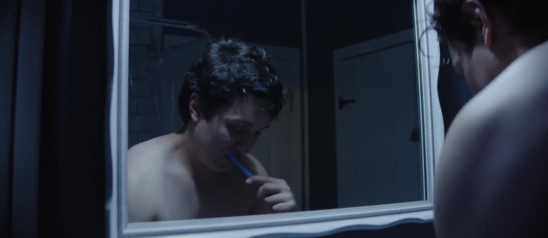 A young man is brushing his teeth in front of a bathroom mirror while his partner passes behind him