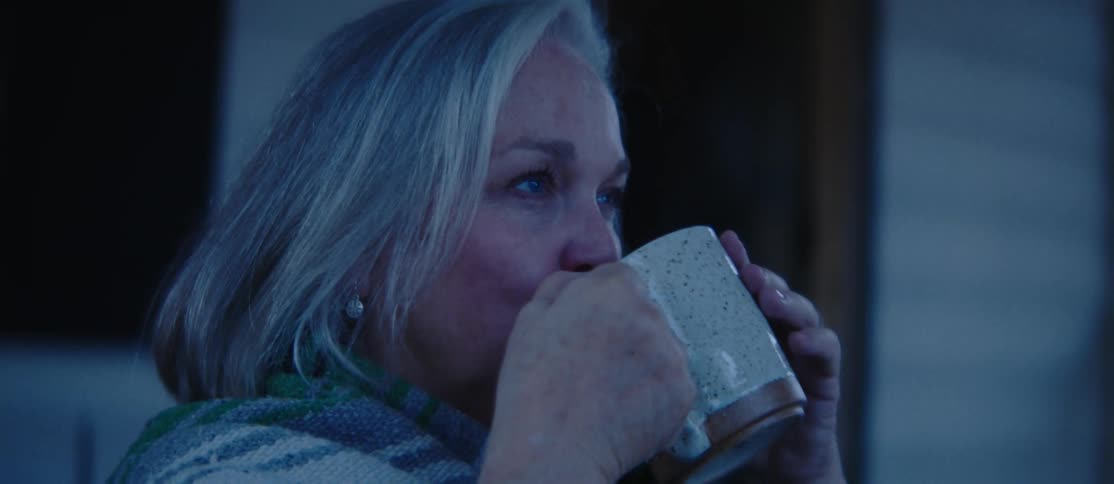 A woman with gray hair is drinking from a speckled mug while wrapped in a striped blanket