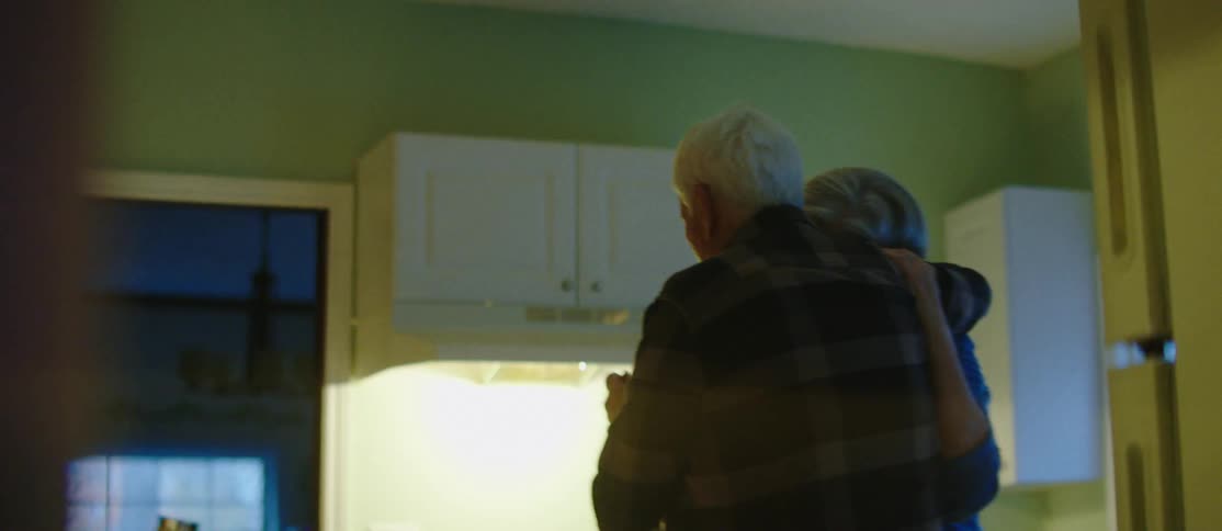 An elderly man and woman are hugging and slowly dancing in a kitchen with white furniture