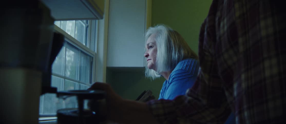 A woman with a head of silver hair and a bright blue shirt gazes out the kitchen window savoring her coffee her husband meanwhile pours a steaming cup and leans in to give her a tender kiss