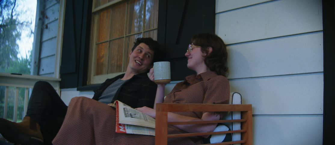 Two people are sitting on a porch one holding a mug and reading a magazine