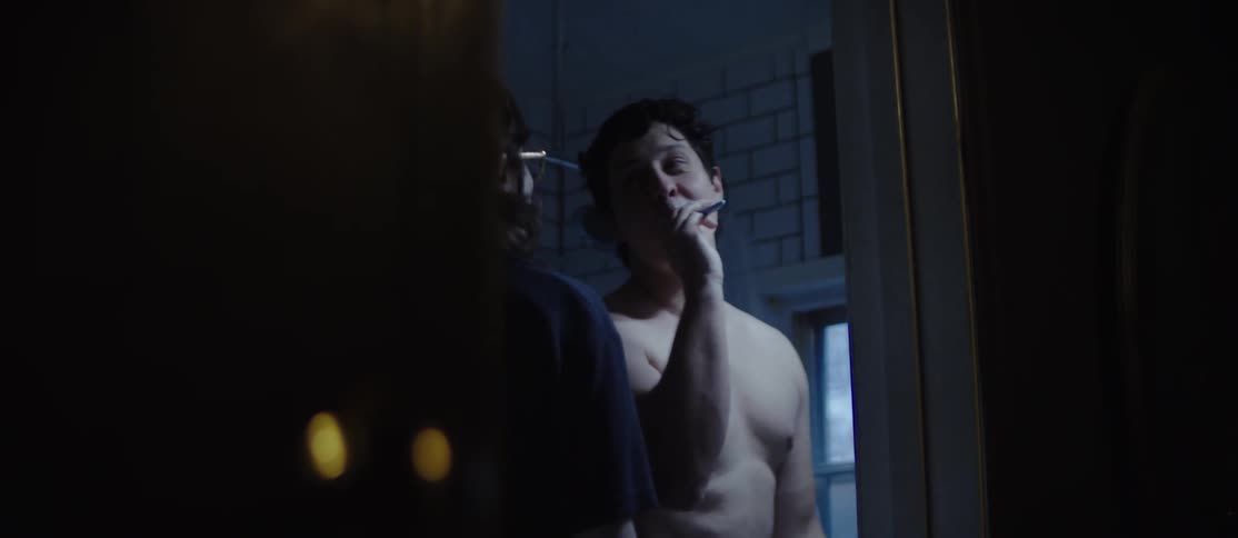 A woman wearing glasses and a black sweater enters the bathroom while a shirtless man brushes his teeth
