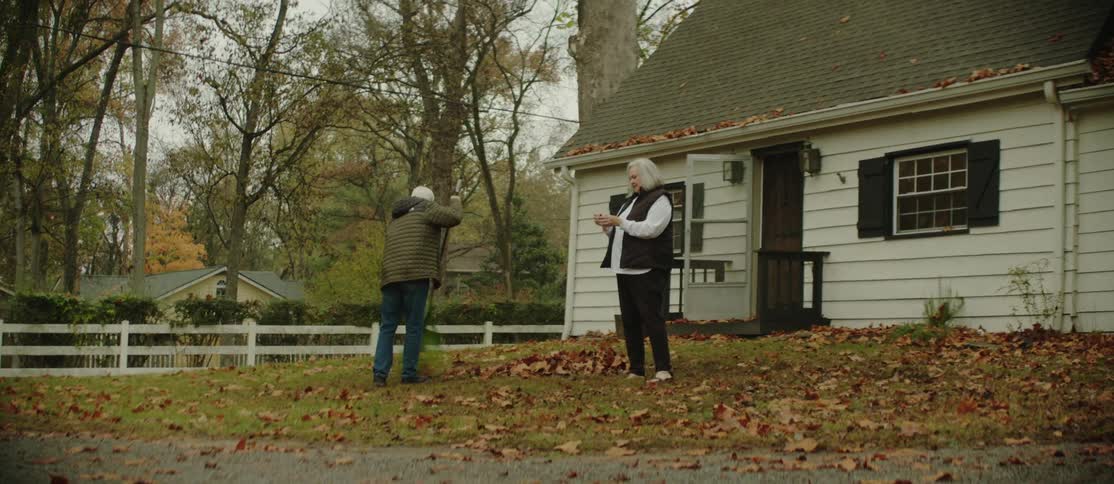 An elderly man rakes leaves from the front porch while his wife inspects them