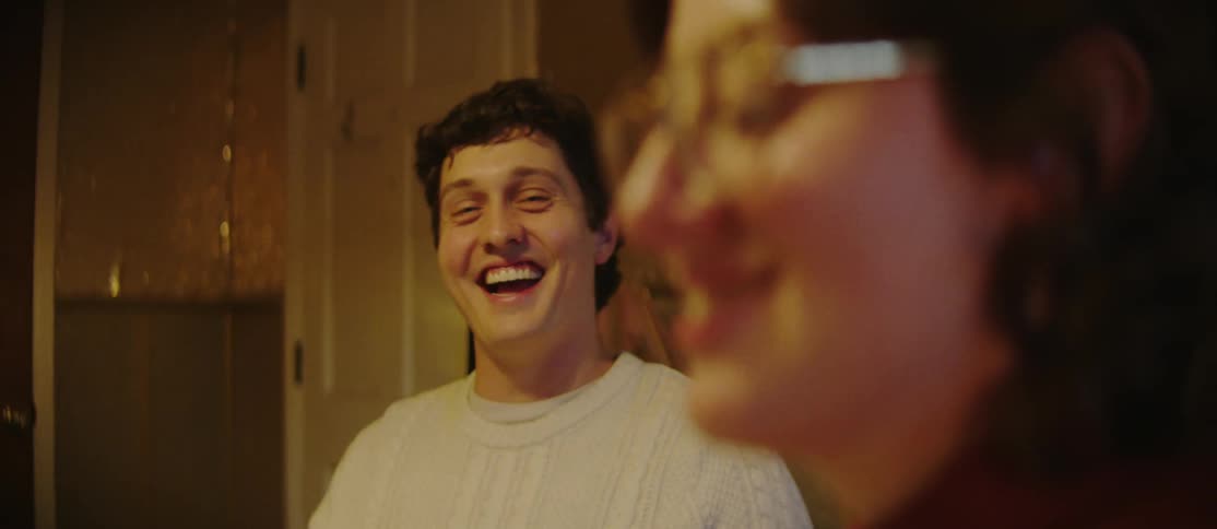A man wearing a white sweater is smiling and laughing while looking at a woman who is talking in front of him