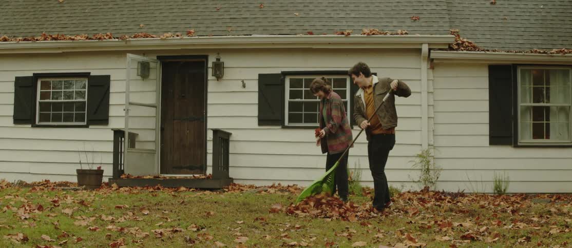 Man and a woman in a backyard cleaning up fallen dry leaves on the grass