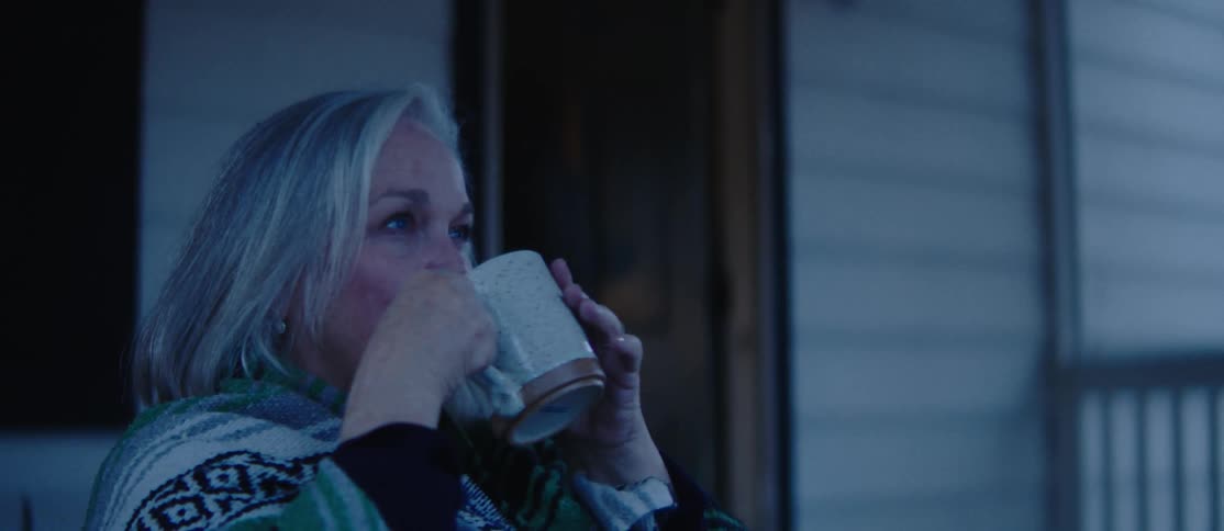 A person is sitting on a porch wrapped in a patterned blanket drinking from a ceramic mug