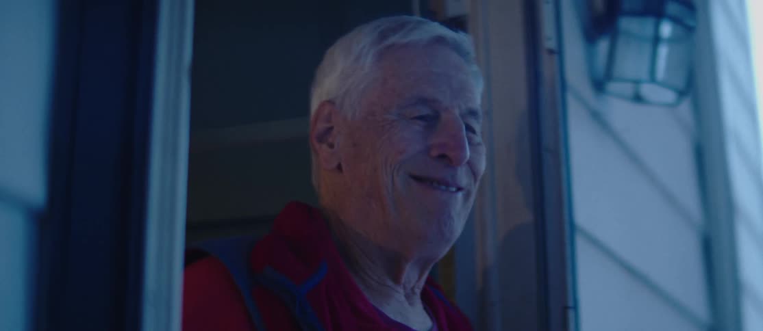 An elderly person with white hair is standing in a doorway drinking from a mug