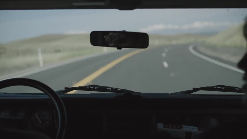 The view of a country road through the windshield of a car with the driver seen in her rearview mirror