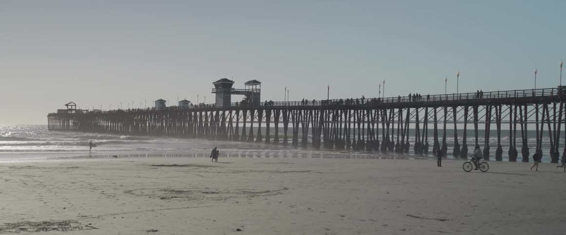 Several people walking on a sandy beach with a huge wooden pier on the right side
