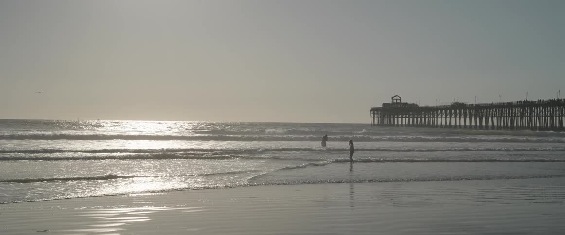 Two people having fun in the sea at the beach with a pier in the background