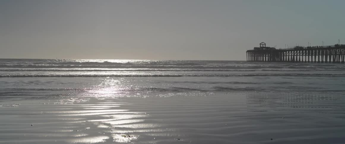 A pier extends into the calm ocean with the sun reflecting off the water and creating a shimmering path across the sand