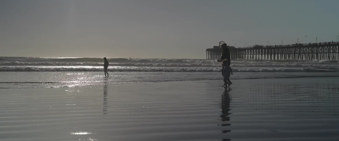 The silhouette of an adult and two children who are walking on the wet sand of a beach near the sea and there is a large pier in the background