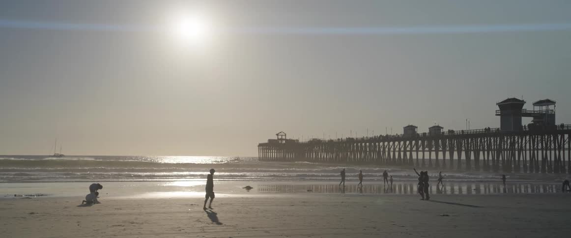 People playing and walking on the sand of the beach with a pier next to it