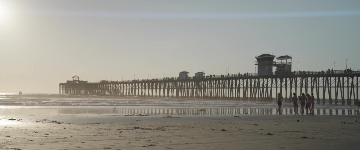 People running and walking on the sand of the beach with a pier in the background