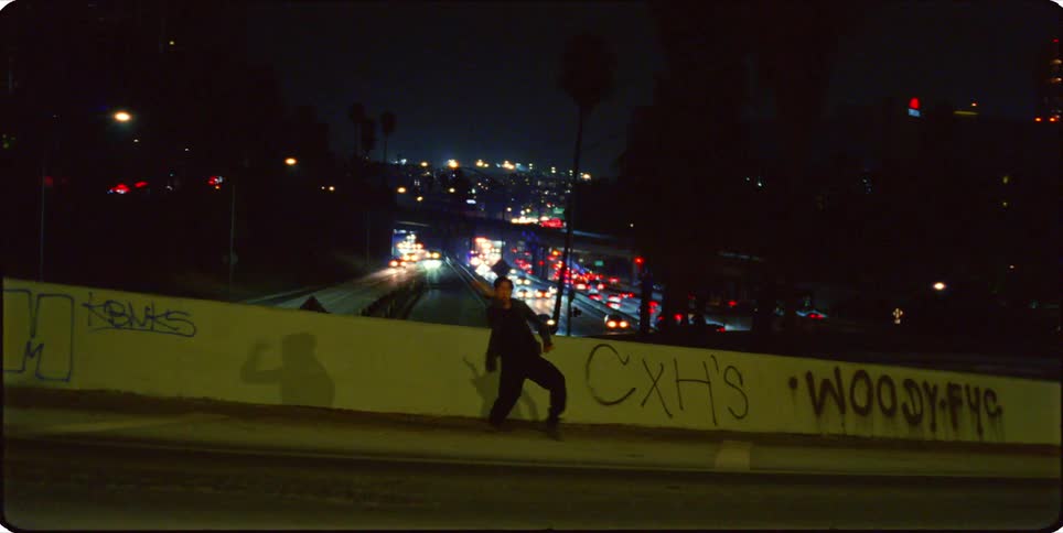 A person is dancing on a wall overlooking a highway with blurred city lights at night