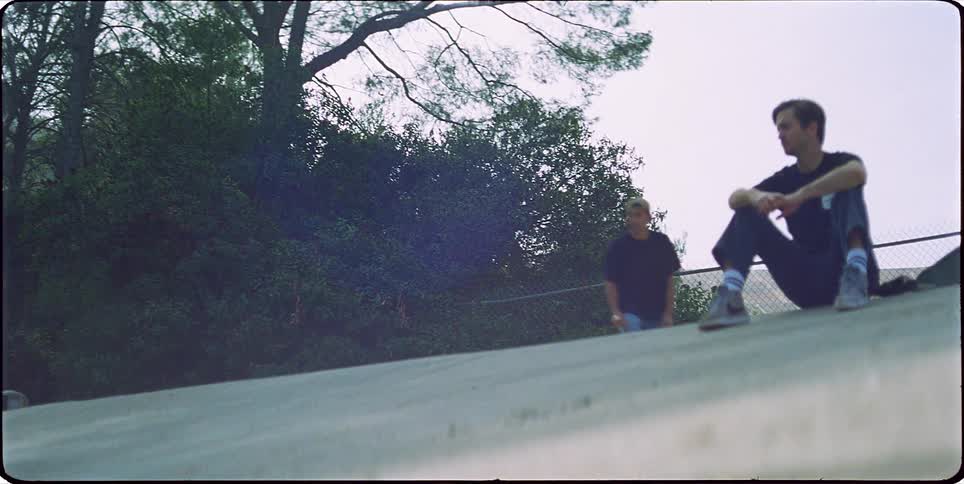 A boy sitting on a concrete ramp and then another boy comes with a skateboard and does a flip and the skateboard falls to the ground