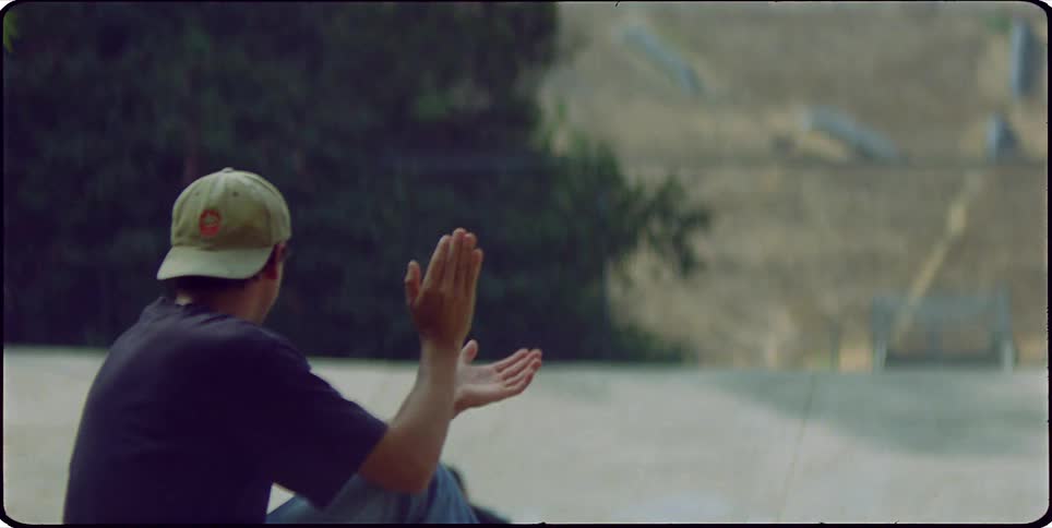 Young man sitting and clapping for a colleague who is doing tricks with a skateboard on an outdoor rink