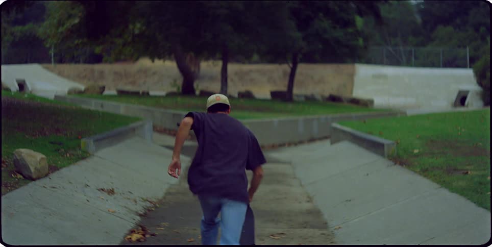 A close up of a young man wearing a baseball cap is followed by a shot of him skateboarding away from the camera down a paved path
