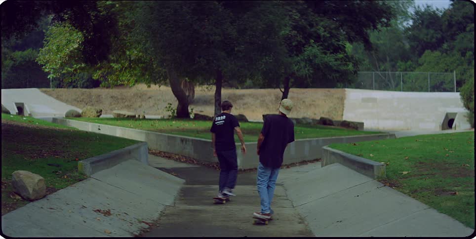 Two skateboarders shake hands then skate away from the camera down a paved path