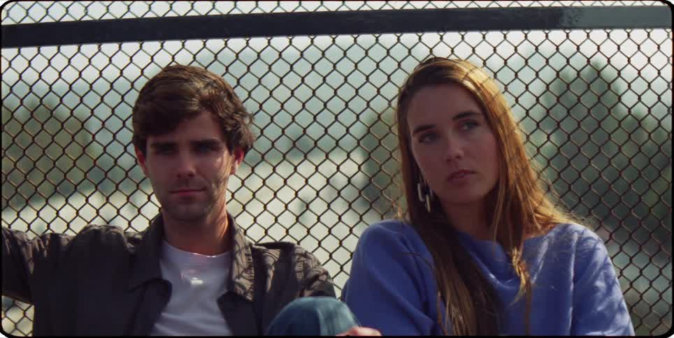 A young man and woman sit together against a chain link fence and the woman rests her head on the mans shoulder and smiles