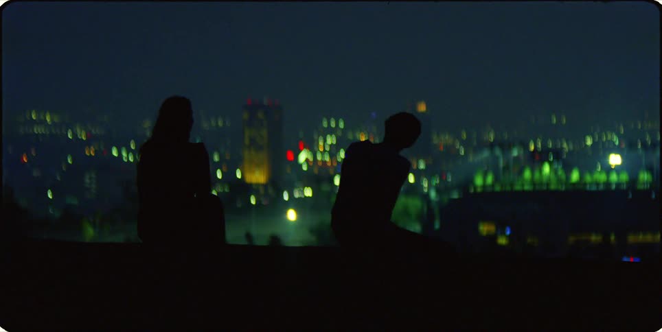 Silhouette of two people sitting outdoors with a panoramic view of an illuminated city at night