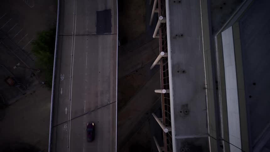 An aerial view shows a motorcycle turning left on a street below an overpass with a black car traveling on the upper level