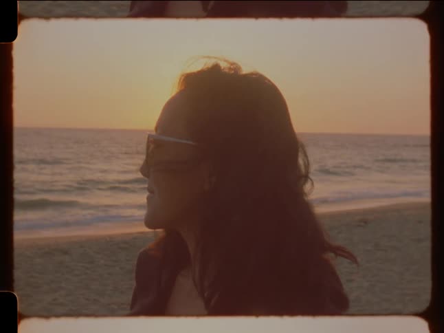 Woman with sunglasses and wavy hair gazes away on the beach at sunset