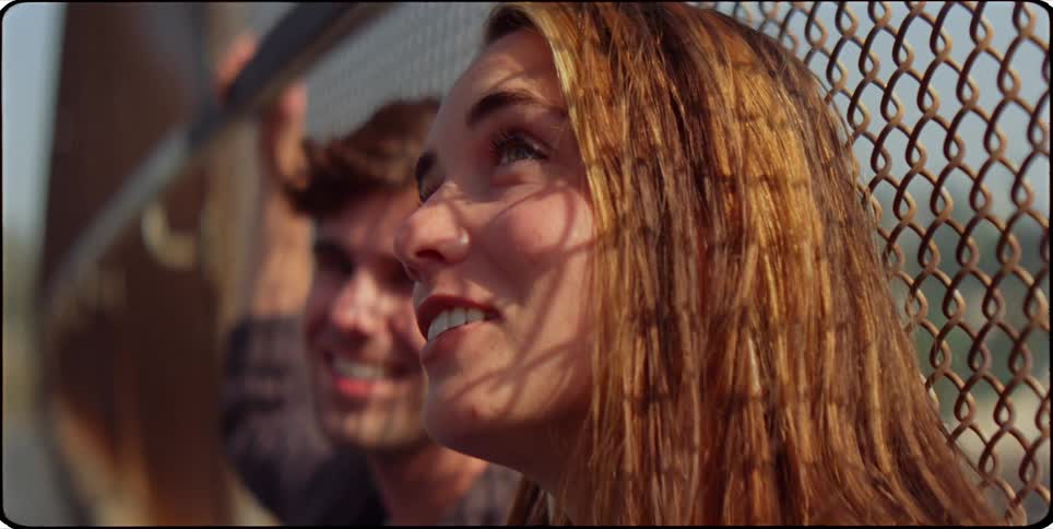 A man and a woman sit on the ground of a fenced pedestrian overpass chatting and laughing