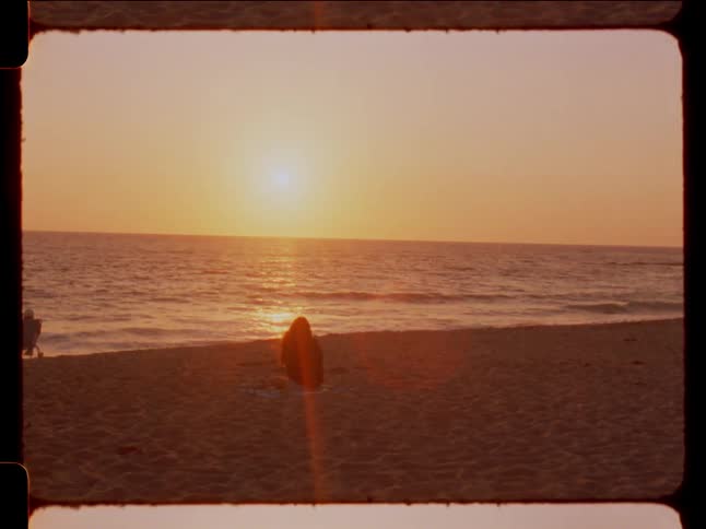 Person sitting on a towel on the sand of the beach facing the sea with the sunset on the horizon