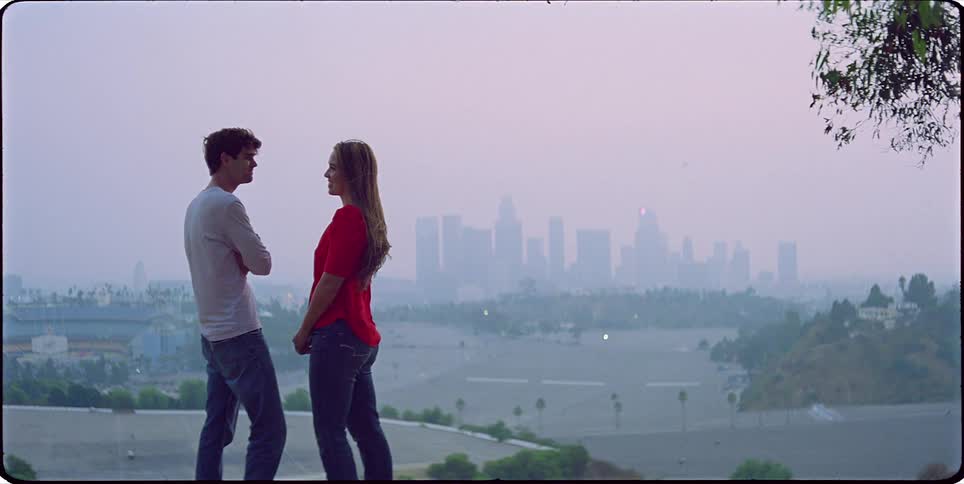 A man wearing a shirt and jeans and a woman with brown hair a red blouse and jeans hug each other with the big city skyline in the background