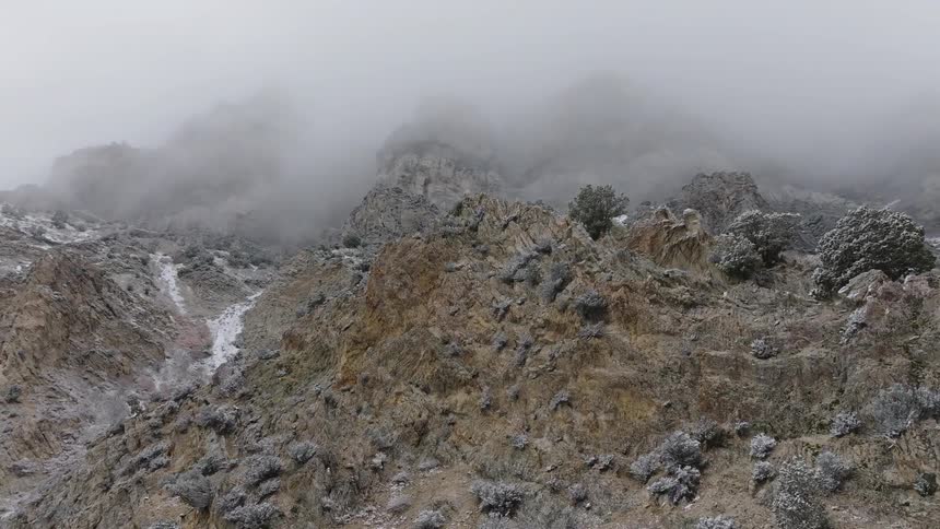 A rugged rocky mountainside dusted with snow is shrouded in fog