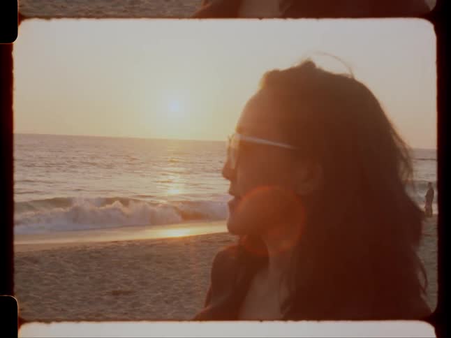 A woman wearing sunglasses looks toward the ocean as the sun sets