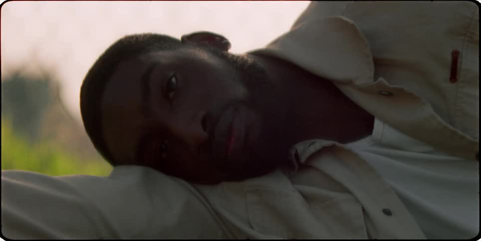 A bearded man wearing a beige jacket and white shirt rests his head on his arm gazing thoughtfully into the distance with a serene outdoor backdrop