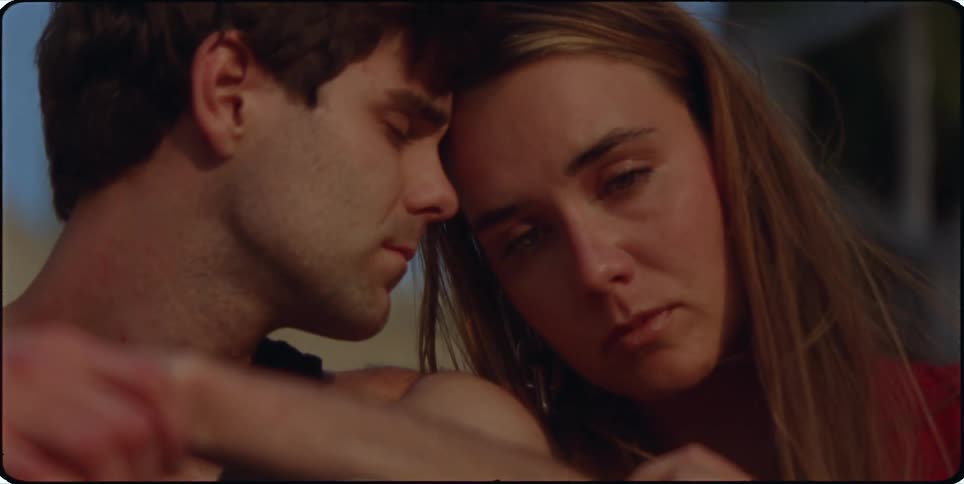 A man and a woman sit closely together leaning their heads against one another sharing a quiet intimate moment with solemn expressions