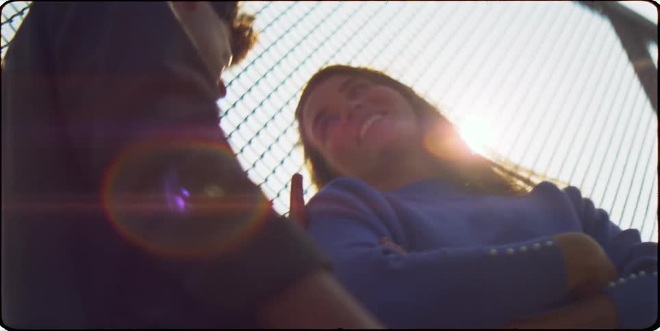 A young man and woman stand face to face and talk smiling and laughing with a chain link fence behind them and the sun shining brightly overhead